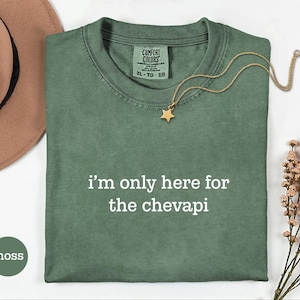 May include: A moss green t-shirt with the text "I'm only here for the chevapi" in white. A gold star pendant necklace rests on the shirt. A brown hat and flowers are in the background.