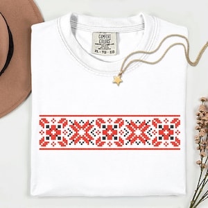 May include: White t-shirt with a red, black, and white geometric embroidered design across the chest. A gold necklace with a star pendant rests on the shirt. The shirt has a Comfort Colors tag.