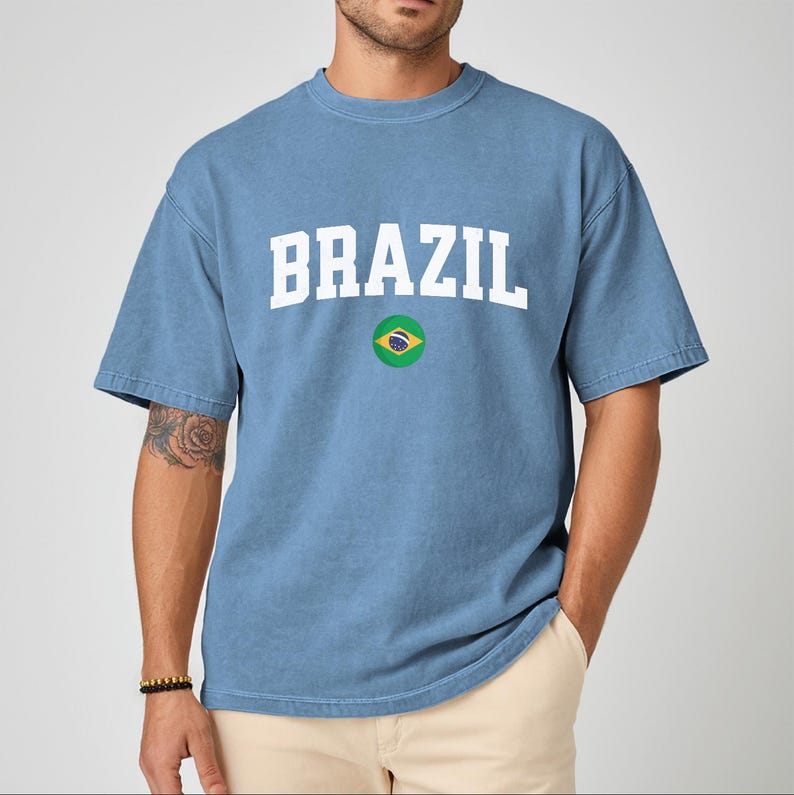 May include: A light blue t-shirt with the word "BRAZIL" in white block letters. Below the word is a circular graphic of the Brazilian flag. The shirt has short sleeves and a crew neck.