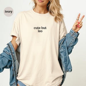 Peut inclure: Un t-shirt &agrave; manches courtes de couleur ivoire avec le texte noir "cute but leo" imprim&eacute; au centre de la poitrine. Le t-shirt pr&eacute;sente une encolure ronde et une coupe d&eacute;contract&eacute;e, port&eacute; sous une veste en jean bleu avec des coutures visibles.