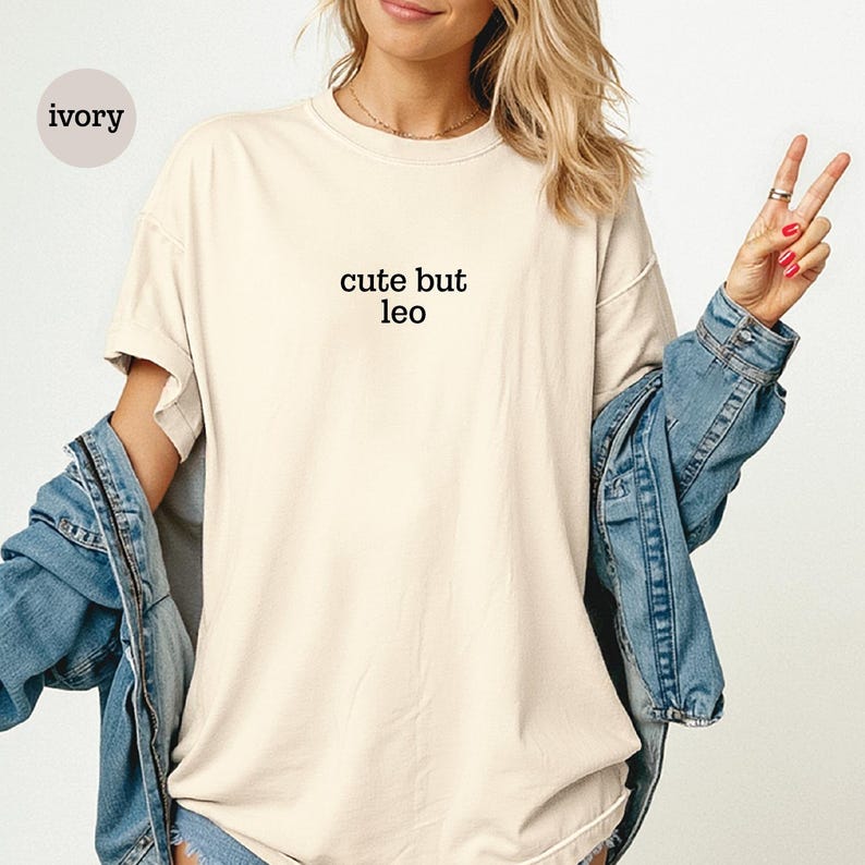 Peut inclure: Un t-shirt &agrave; manches courtes de couleur ivoire avec le texte noir "cute but leo" imprim&eacute; au centre de la poitrine. Le t-shirt pr&eacute;sente une encolure ronde et une coupe d&eacute;contract&eacute;e, port&eacute; sous une veste en jean bleu avec des coutures visibles.