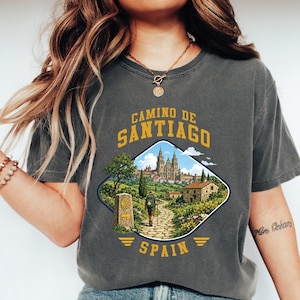 May include: Dark gray t-shirt with a graphic of the Camino de Santiago in Spain. The graphic features a pilgrim walking a path towards a cathedral, with the words "Camino de Santiago" and "Spain" in gold lettering.