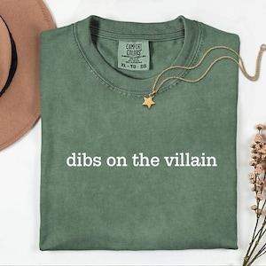May include: A sage green short-sleeve t-shirt with the text "dibs on the villain" printed in white across the chest. The shirt features a Comfort Colors brand label at the inner neckline, indicating a size XL, and is made of 100% cotton fabric.