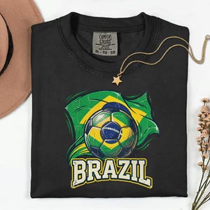 Pode incluir: Camiseta preta com uma bola de futebol e o design da bandeira brasileira. A palavra "BRAZIL" &eacute; impressa em uma fonte vintage. Um colar com pingente de estrela dourada est&aacute; drapeado sobre a camiseta. A camiseta tem uma etiqueta Comfort Colors.