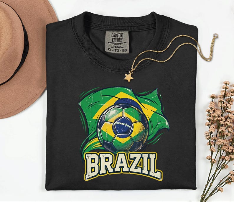 Pode incluir: Camiseta preta com uma bola de futebol e o design da bandeira brasileira. A palavra "BRAZIL" &eacute; impressa em uma fonte vintage. Um colar com pingente de estrela dourada est&aacute; drapeado sobre a camiseta. A camiseta tem uma etiqueta Comfort Colors.