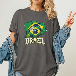Pode incluir: Camiseta cinza escuro com uma bola de futebol e a bandeira brasileira. A palavra "BRAZIL" &eacute; impressa em amarelo e branco abaixo do gr&aacute;fico. A camisa tem mangas curtas e um corte folgado.