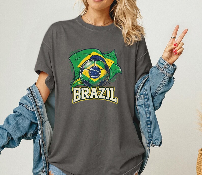 Pode incluir: Camiseta cinza escuro com uma bola de futebol e a bandeira brasileira. A palavra "BRAZIL" &eacute; impressa em amarelo e branco abaixo do gr&aacute;fico. A camisa tem mangas curtas e um corte folgado.