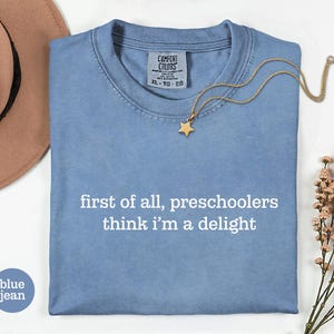 May include: A blue jean coloured t-shirt with the white text "first of all, preschoolers think i'm a delight" printed on the front. The shirt features a Comfort Colors brand label at the inner neckline with size XL-TG-EG clearly visible.