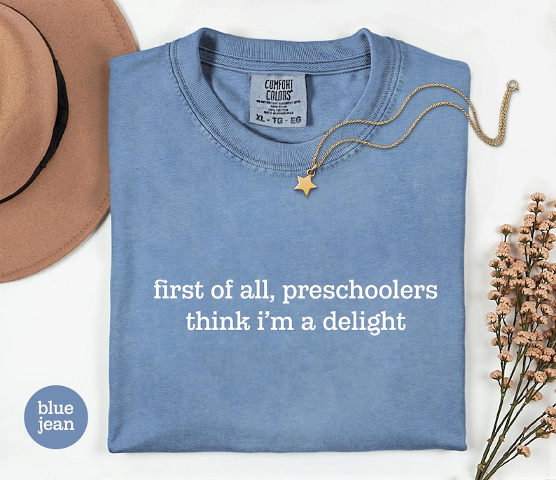 May include: A blue jean coloured t-shirt with the white text "first of all, preschoolers think i'm a delight" printed on the front. The shirt features a Comfort Colors brand label at the inner neckline with size XL-TG-EG clearly visible.