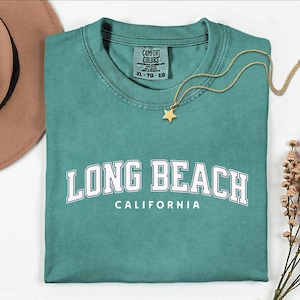 May include: Teal-colored t-shirt with the words "LONG BEACH CALIFORNIA" in white block letters. A gold star pendant necklace rests on the shirt. A brown hat and sprigs of dried flowers are in the background.