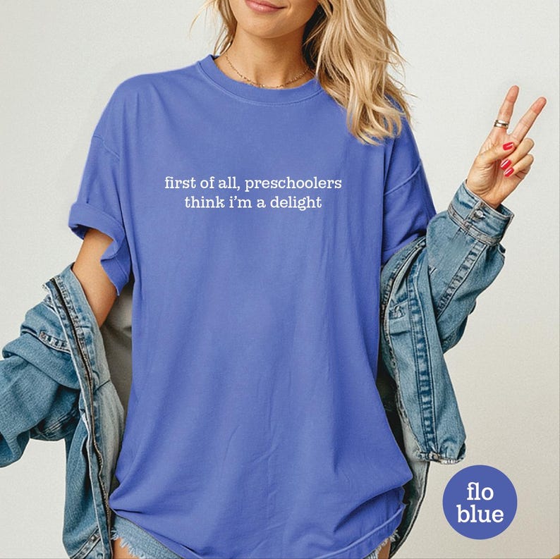 May include: A periwinkle blue short-sleeved t-shirt featuring the white text "first of all, preschoolers think i'm a delight" printed on the chest. The shirt has a crew neckline and a relaxed fit, paired with a blue denim jacket draped over the shoulders.