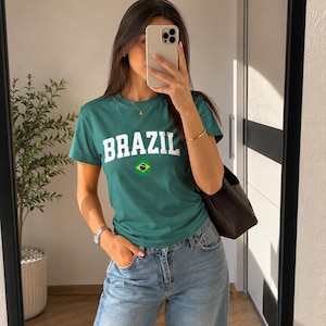 May include: Green t-shirt with the word "BRAZIL" in white block letters and a small Brazilian flag graphic. The shirt is paired with blue jeans.
