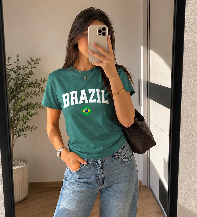 May include: Green t-shirt with the word "BRAZIL" in white block letters and a small Brazilian flag graphic. The shirt is paired with blue jeans.