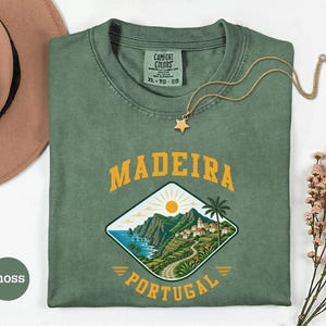 May include: A moss green t-shirt with a graphic of Madeira, Portugal. The design features a coastal landscape with mountains, a road, and the text "MADEIRA PORTUGAL" in yellow. A gold necklace with a star pendant rests on the shirt.