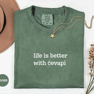 Peut inclure: Un t-shirt à manches courtes vert mousse avec un col rond et le texte blanc "life is better with ćevapi" imprimé sur la poitrine. Le t-shirt présente une étiquette de marque Comfort Colors au niveau du col intérieur, indiquant une taille XL.