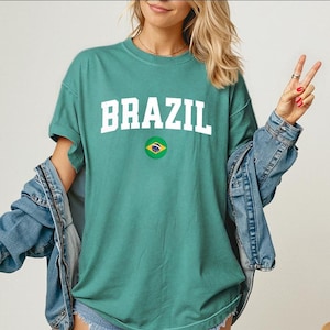 May include: A teal t-shirt with the word "BRAZIL" in white block letters and a small Brazilian flag emblem. The shirt is paired with a denim jacket.