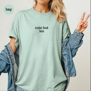 Peut inclure: Un t-shirt &agrave; manches courtes vert clair avec le texte noir "cute but leo" imprim&eacute; au centre de la poitrine. Le t-shirt pr&eacute;sente une encolure ronde et une coupe d&eacute;contract&eacute;e, port&eacute; sous une veste en jean bleue &agrave; manches longues.