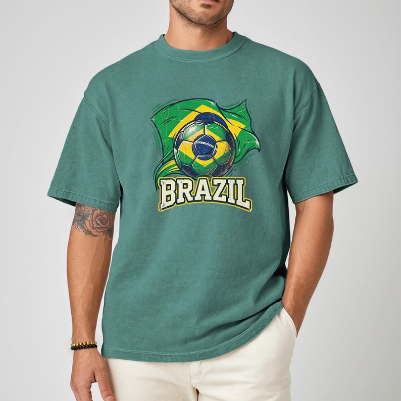 Pode incluir: Camiseta verde com uma bola de futebol e o design da bandeira brasileira. A palavra "BRAZIL" &eacute; impressa em fonte amarela e branca abaixo da bola de futebol. A camiseta &eacute; de cor verde lisa.