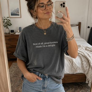 May include: A charcoal grey short-sleeved t-shirt featuring the white text "first of all, preschoolers think i'm a delight" printed on the chest. The shirt has a crew neckline and a relaxed fit, paired with light blue denim jeans.