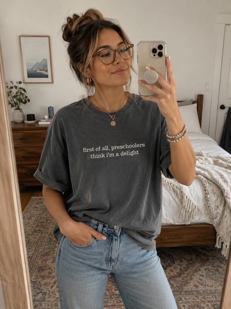 May include: A charcoal grey short-sleeved t-shirt featuring the white text "first of all, preschoolers think i'm a delight" printed on the chest. The shirt has a crew neckline and a relaxed fit, paired with light blue denim jeans.