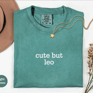 Peut inclure: Un t-shirt &agrave; manches courtes vert sarcelle avec le texte "cute but leo" imprim&eacute; en blanc sur la poitrine. Le t-shirt pr&eacute;sente une &eacute;tiquette de marque Comfort Colors &agrave; l'encolure int&eacute;rieure. Un collier dor&eacute; avec un petit pendentif &eacute;toile repose sur le tissu.