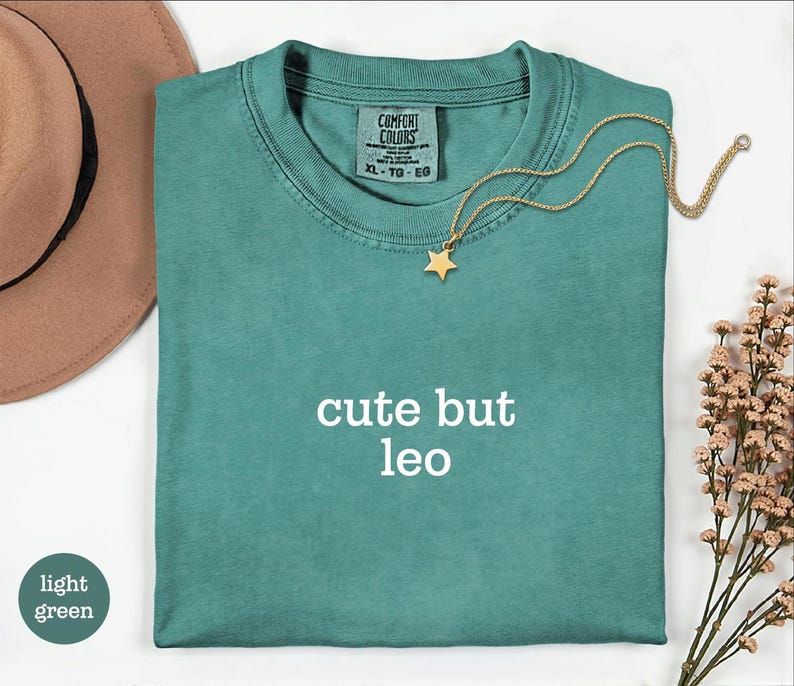 Peut inclure: Un t-shirt &agrave; manches courtes vert sarcelle avec le texte "cute but leo" imprim&eacute; en blanc sur la poitrine. Le t-shirt pr&eacute;sente une &eacute;tiquette de marque Comfort Colors &agrave; l'encolure int&eacute;rieure. Un collier dor&eacute; avec un petit pendentif &eacute;toile repose sur le tissu.