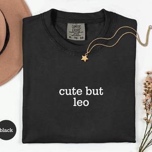 Peut inclure: Un t-shirt noir avec le texte blanc 'cute but leo' imprim&eacute; sur le devant. Le t-shirt pr&eacute;sente une &eacute;tiquette Comfort Colors au niveau du col indiquant une taille XL. Un collier dor&eacute; avec un petit pendentif en forme d'&eacute;toile repose sur le tissu.