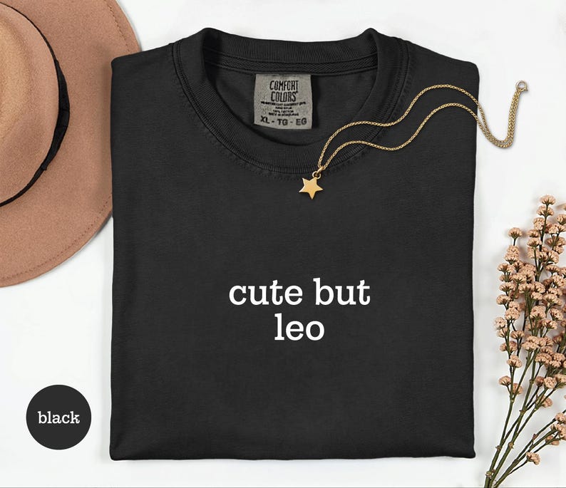 Peut inclure: Un t-shirt noir avec le texte blanc 'cute but leo' imprim&eacute; sur le devant. Le t-shirt pr&eacute;sente une &eacute;tiquette Comfort Colors au niveau du col indiquant une taille XL. Un collier dor&eacute; avec un petit pendentif en forme d'&eacute;toile repose sur le tissu.