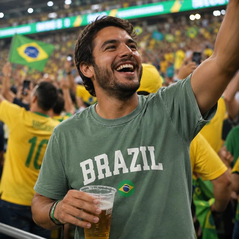 May include: A person wearing a green t-shirt with the word "BRAZIL" in white letters and a small Brazilian flag graphic. The person is smiling and holding a clear plastic cup with a light-colored beverage. The background shows a crowd of people.