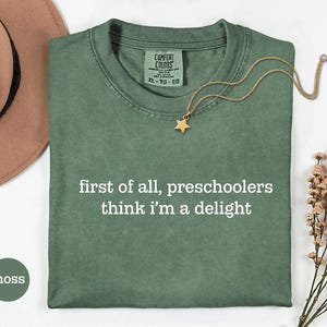 May include: A moss green short-sleeved t-shirt with a crew neckline and the white printed text "first of all, preschoolers think i'm a delight" centred on the chest. The shirt features a Comfort Colors brand label at the inner neck area.