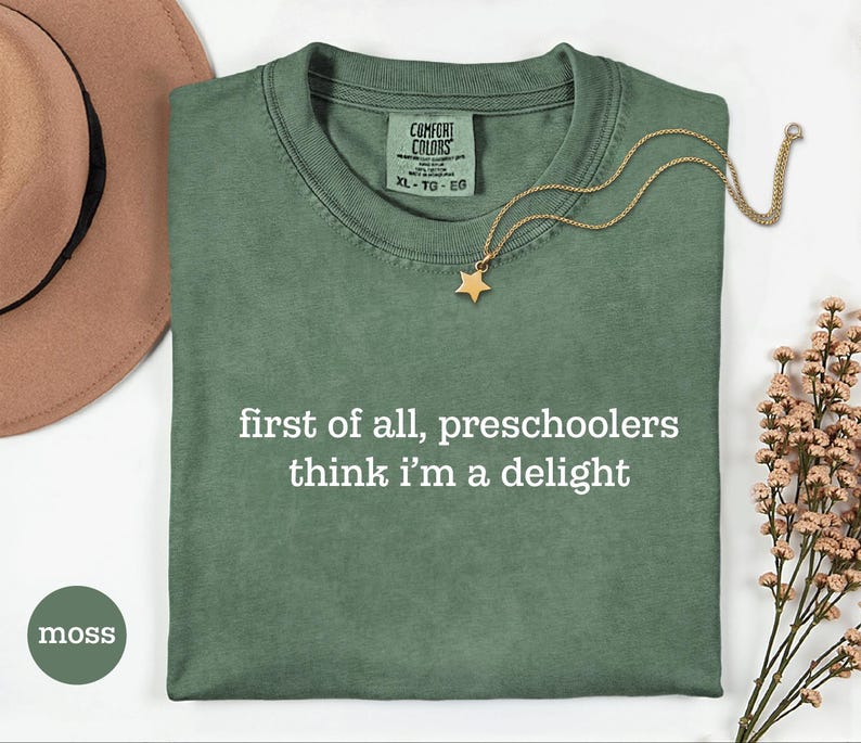 May include: A moss green short-sleeved t-shirt with a crew neckline and the white printed text "first of all, preschoolers think i'm a delight" centred on the chest. The shirt features a Comfort Colors brand label at the inner neck area.