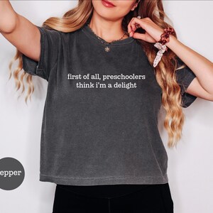 May include: A charcoal grey short-sleeve t-shirt with a crew neckline. The shirt features white text printed on the chest that reads, first of all, preschoolers think i'm a delight. A small circular label in the bottom left corner says pepper.