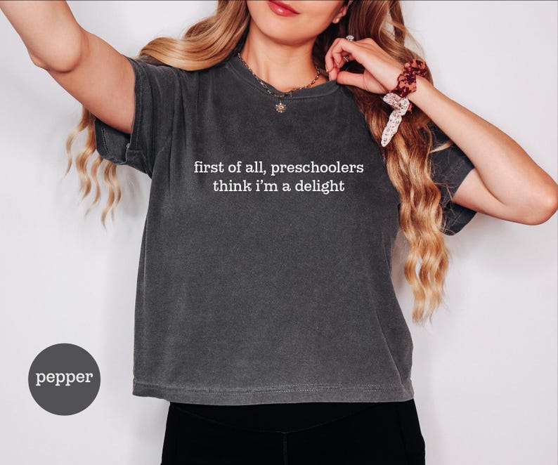 May include: A charcoal grey short-sleeve t-shirt with a crew neckline. The shirt features white text printed on the chest that reads, first of all, preschoolers think i'm a delight. A small circular label in the bottom left corner says pepper.