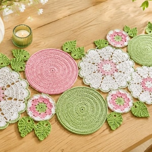 Op de afbeelding: Een handgemaakte gehaakte tafelloper met een bloemmotief in wit, roze en groen. De loper bevat ronde placemats en bladeraccenten, gerangschikt op een houten tafel. Een vaas met bloemen en een kaars zijn ook aanwezig.