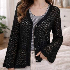 Könnte beinhalten: Ein schwarzer Cardigan mit offenem Webmuster, V-Ausschnitt und Knopfverschluss. Der Cardigan hat lange, ausgestellte Ärmel und wird über einem gestreiften Tanktop und hellen Hosen getragen. Der Gesamtstil ist lässig und feminin.