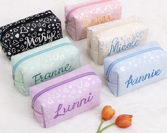 Könnte beinhalten: Eine Sammlung personalisierter Kosmetiktaschen in verschiedenen Pastellfarben, darunter Schwarz, Rosa, Mintgrün, Creme, Hellviolett und Blau. Jede Tasche hat einen Namen in einer passenden Farbe und ein Herzmuster.