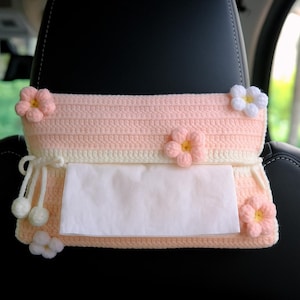Könnte beinhalten: Ein gehäkelter Taschentuchhalter in Pfirsich- und Cremefarben mit Blumenakzenten. Der Halter ist für die Aufhängung an einer Kopfstütze im Auto konzipiert. Die Blumen sind pfirsichfarben und weiß und verleihen dem Artikel eine dekorative Note.