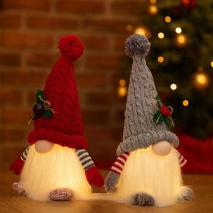 May include: Two Christmas gnome decorations. One gnome has a red knit hat with a pom-pom, the other has a gray knit hat with a pom-pom. Both have white beards, striped arms, and glowing bodies. Each gnome is decorated with a sprig of holly.