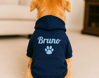 May include: A golden retriever wearing a navy blue dog hoodie. The hoodie features the name "Bruno" in a script font above a light blue paw print. The dog is sitting on a light-colored surface.