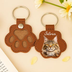 May include: Two brown leather paw-shaped keyrings with silver rings. One has a paw print cutout, the other features a cat portrait and the name "Silvia" in white. The keyrings are on a light background.