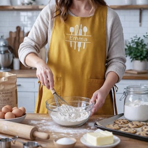 May include: Mustard yellow apron with white kitchen utensil graphics and the name "Emma". The apron is worn while mixing ingredients in a glass bowl. Baking supplies include flour, eggs, butter, and a tray of cookies.