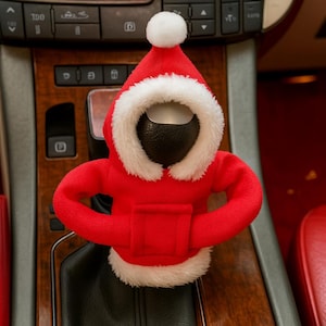 Op de afbeelding: Een rode kerstman-themaversnellingspookhoes voor auto's met witte imitatiebontafwerking en een witte pompon op de hoed. De hoes is ontworpen om over de versnellingspook van een auto te passen en een feestelijke touch toe te voegen.