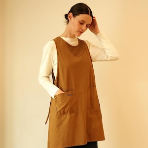May include: A brown apron with two large front pockets and the word "CONTAIN" embroidered on the left pocket. The apron is worn over a white long-sleeved shirt. The apron has a sleeveless design and a tie on the side.
