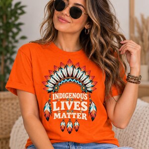 Può includere: Una t-shirt arancione a maniche corte con una stampa grafica di un copricapo di piume colorate sopra il testo "INDIGENOUS LIVES MATTER" in lettere maiuscole bianche, accentuato da ulteriori illustrazioni di piume e piccole perline.