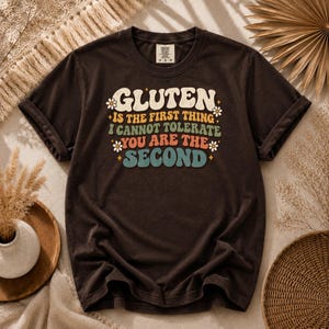 Può includere: Una t-shirt a maniche corte marrone scuro con il testo "GLUTEN IS THE FIRST THING I CANNOT TOLERATE YOU ARE THE SECOND" stampato al centro del petto in un carattere multicolore con piccoli accenti a forma di fiore e stella.