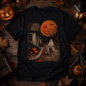 Può includere: Una t-shirt nera con una stampa grafica che raffigura una figura avvolta in un lenzuolo bianco in bicicletta lungo una strada di notte. La scena include una grande luna piena arancione, alberi spogli, pipistrelli e zucche a terra.