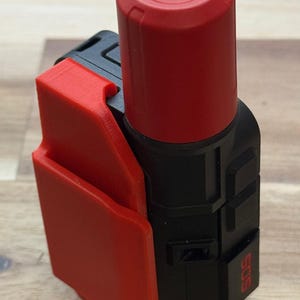 Può includere: Un dispositivo nero e rosso con una parte superiore cilindrica e un pannello laterale rettangolare. La parte superiore è rossa e il pannello laterale è anch'esso rosso. Il dispositivo ha le lettere "SOS" stampate sul lato. È probabilmente uno strumento di sicurezza o di emergenza.