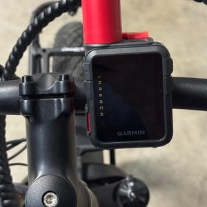 Puede incluir: Un dispositivo GPS Garmin InReach negro montado en un manillar de bicicleta. El dispositivo tiene una parte superior roja y la palabra "GARMIN" es visible. El manillar y la potencia son negros.