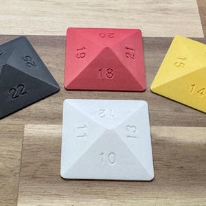 Puede incluir: Cuatro dados geométricos en gris, rojo, blanco y amarillo. Cada dado tiene forma de pirámide con números en cada cara. Los dados están dispuestos sobre una superficie de madera, adecuados para juegos de mesa.
