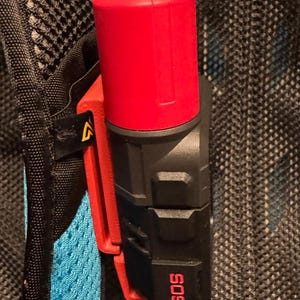 May include: A black and red emergency device attached to a backpack. The device has a red top and a black body with the letters "SOS" in white. The device is clipped to a red and black clip.